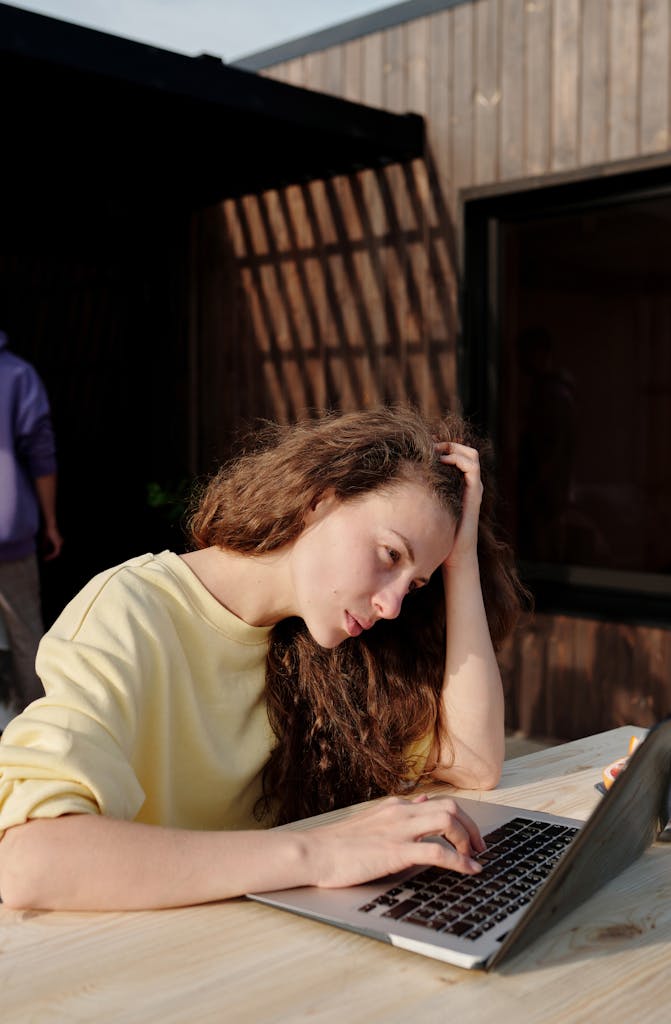 A young woman with long hair works outdoors on a laptop, appearing focused and thoughtful.