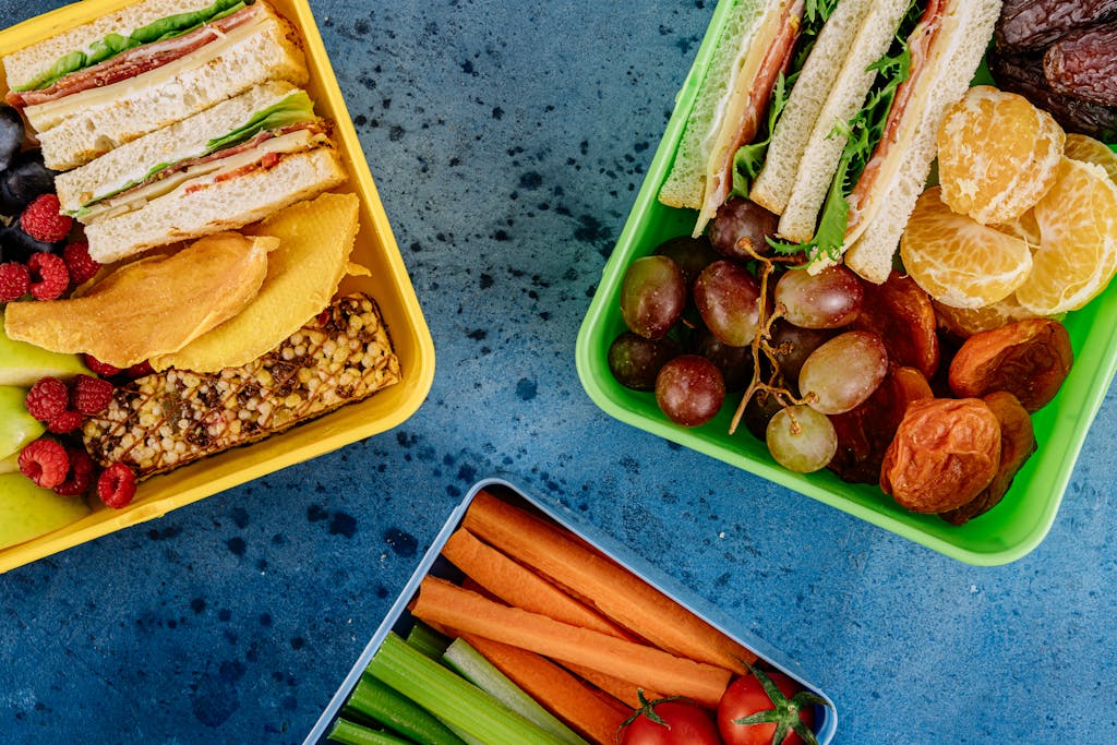 Top view of vibrant lunch boxes filled with sandwiches, fruits, and vegetables on a blue surface.