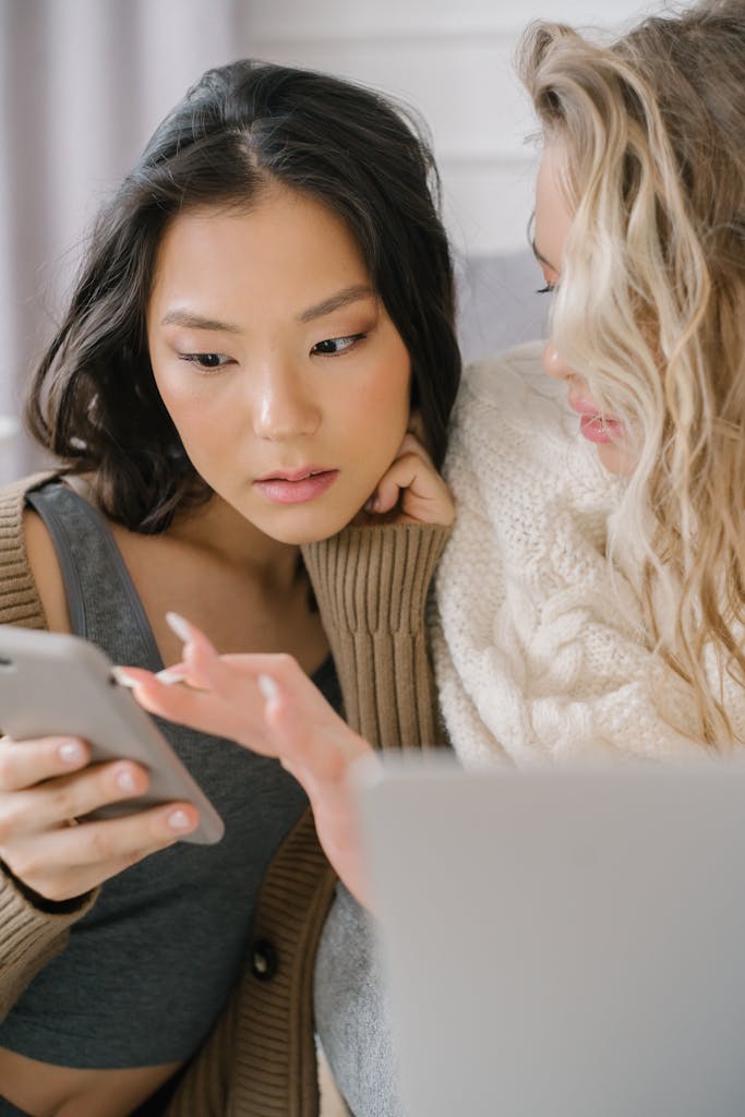 Two women engrossed in a mobile device while sitting indoors, showcasing friendship.