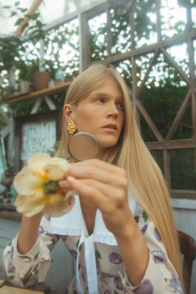 Woman in a floral dress with a magnifying glass inspecting a flower inside a greenhouse.
