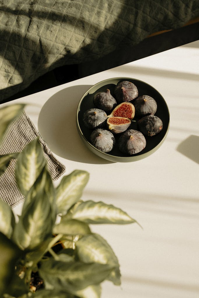 A bowl of fresh figs on a sunlit table with green foliage for a serene food photography setting.