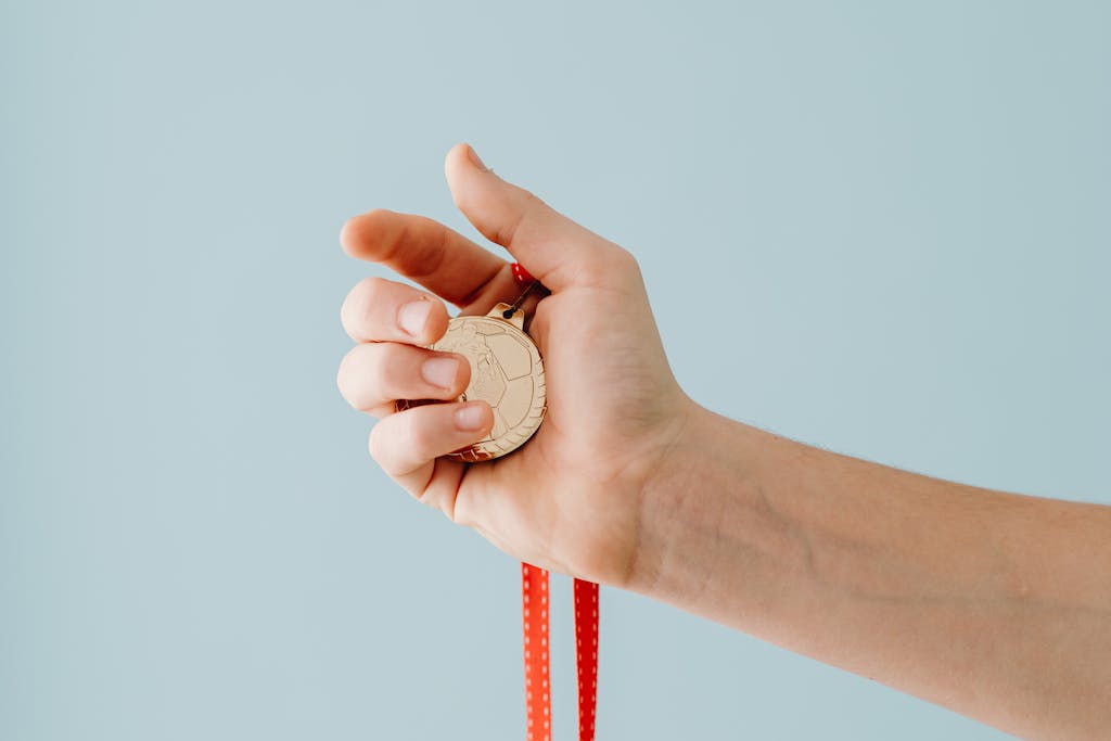 A close-up image of a hand holding a gold medal with a red ribbon against a blue background.