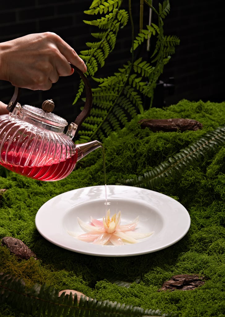 A delicate glass teapot pours vibrant tea over a flower on a mossy bed, showcasing nature's beauty.