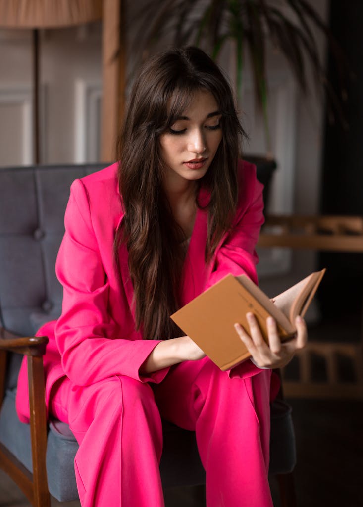 A fashionable woman wearing a pink suit reads a book indoors, embodying modern style.