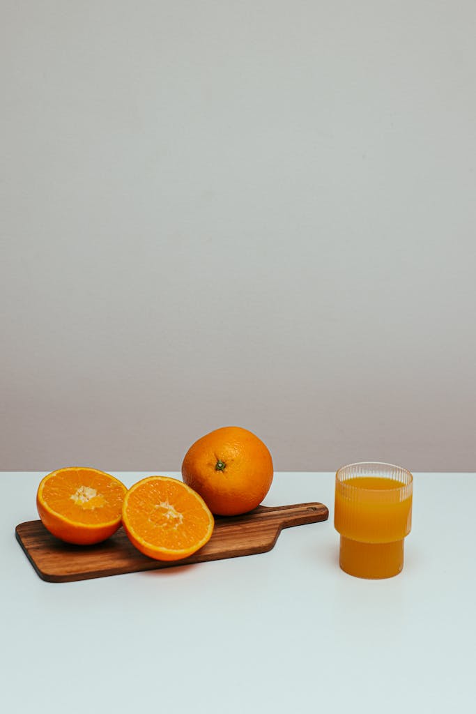 A fresh glass of orange juice with sliced oranges on a wooden board against a minimalist background.