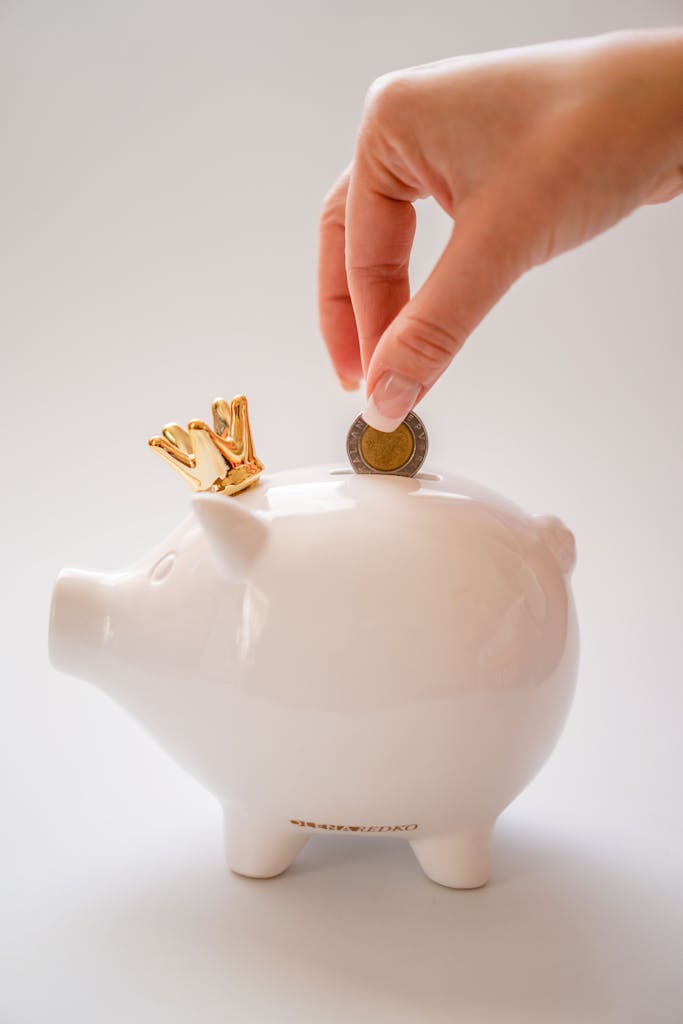 A hand deposits a coin into a white piggy bank with a golden crown, symbolizing savings.