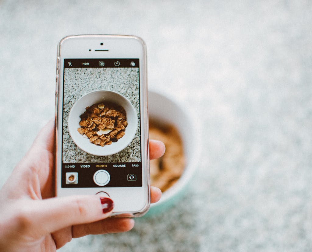 A hand holding a smartphone taking a photo of a bowl of cereal.