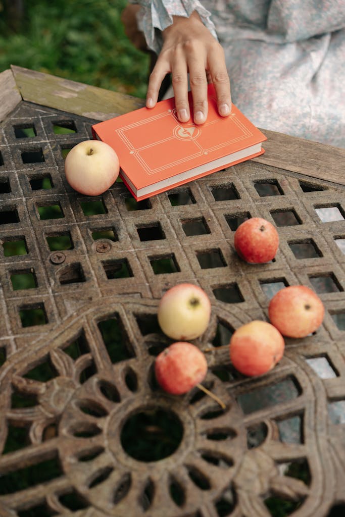 A hand reaches for an orange book surrounded by apples on an outdoor metal table.
