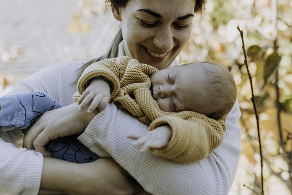 A joyful mother in autumn attire proudly holds her sleeping baby in a peaceful outdoor setting.