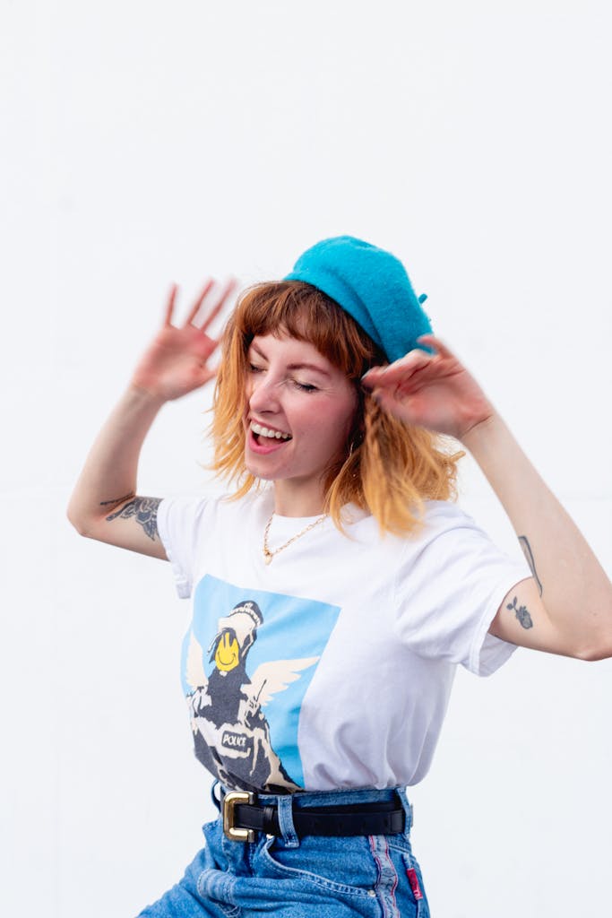 A lively young woman with tattoos dances joyfully in casual fashion and a blue hat.