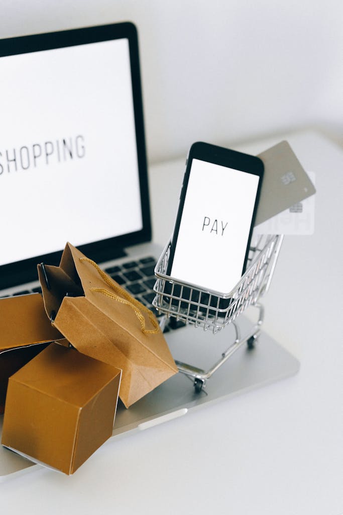 A miniature shopping cart holds a smartphone and credit card on a laptop, symbolizing online shopping.