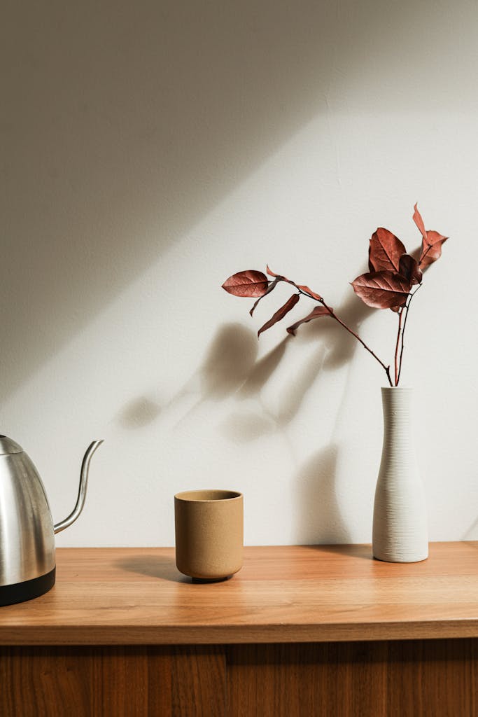 A minimalist interior setup featuring a dried leaf arrangement in a white ceramic vase on a wooden cabinet.