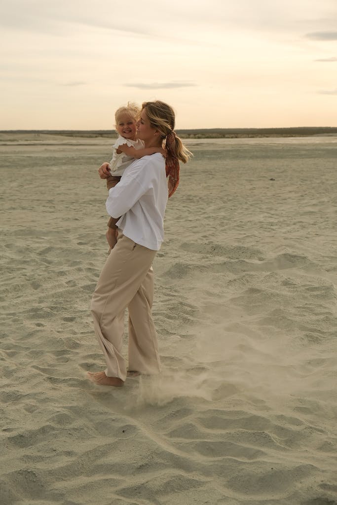 A mother and baby walking on a sandy beach during sunset, sharing a joyful moment.