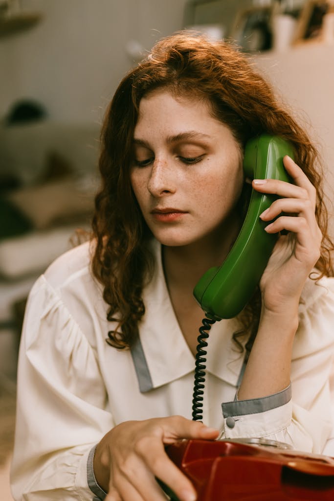 A pensive redhead woman uses a vintage green telephone, creating a nostalgic indoor scene.