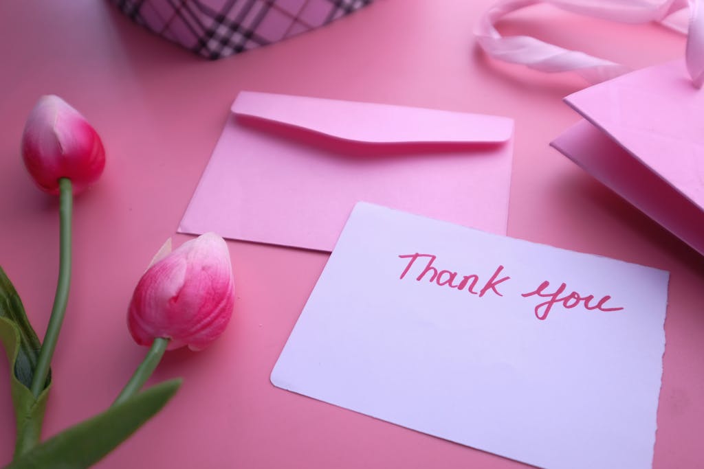 A pink thank you card with flowers and envelope on a pastel surface.