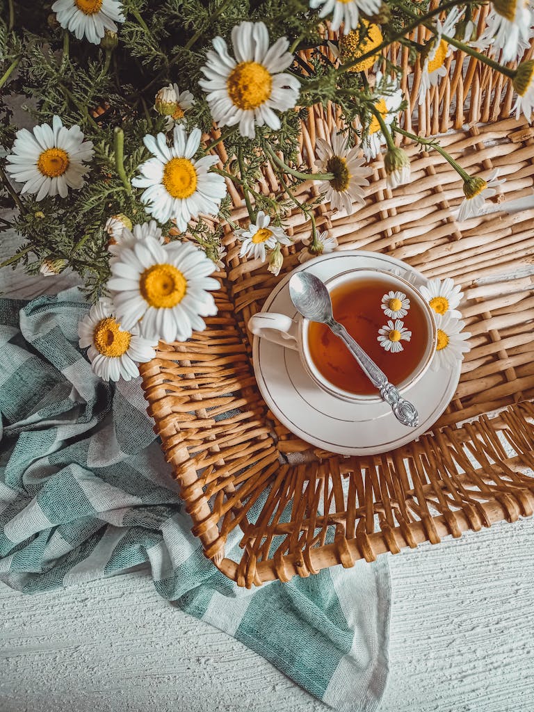 A serene flat lay of camomile tea in a ceramic cup with flowers on a woven tray.