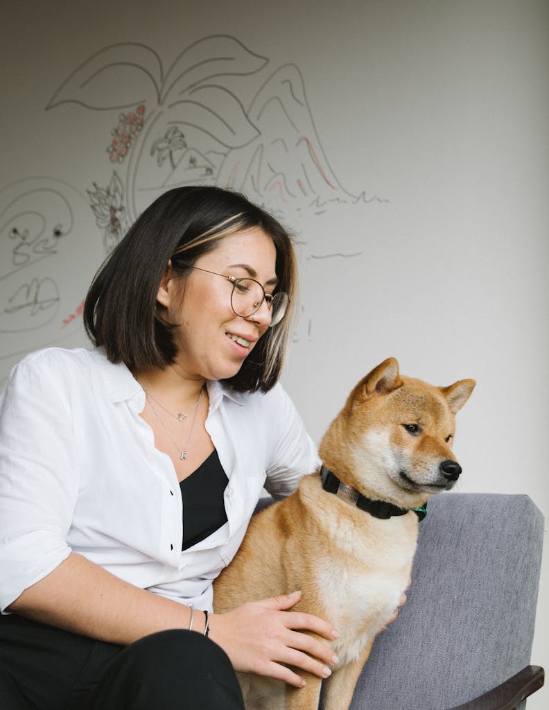 A smiling woman in glasses sits with her Shiba Inu dog, enjoying a cozy moment indoors.