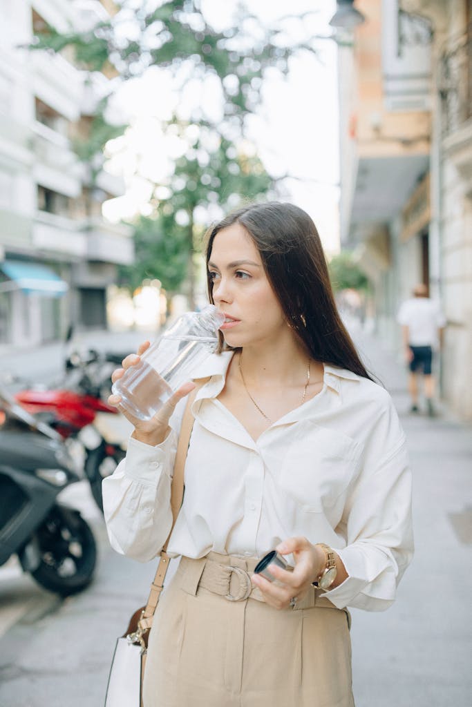 A stylish woman in white shirt sipping water on a sunny city street