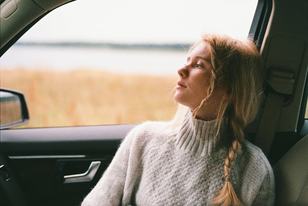 A thoughtful woman with braided hair gazes out a car window during a daytime road trip.