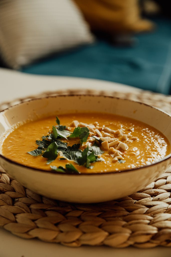 A warm bowl of pumpkin soup garnished with herbs and nuts on a woven mat.