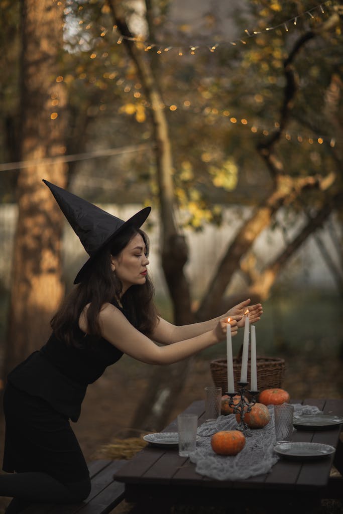 A woman dressed as a witch arranging candles on a table decorated with pumpkins outdoors.