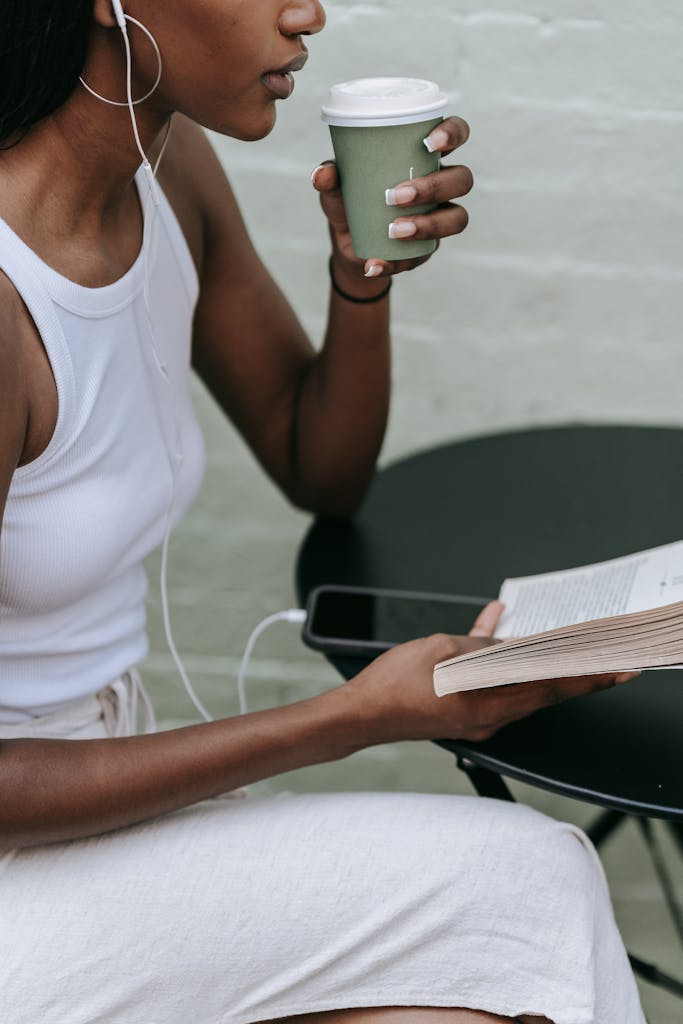 A woman enjoys reading and sipping coffee at an outdoor cafe table, wearing headphones.
