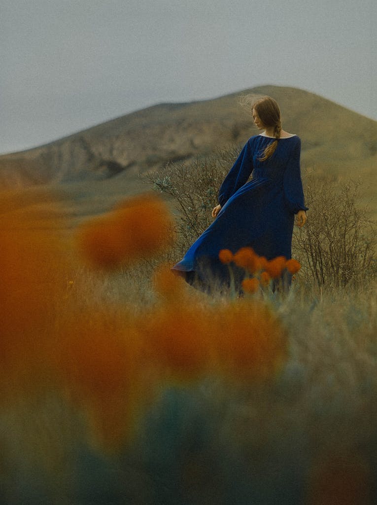 A woman in a blue dress walks through a meadow with flowers, set against a mountain backdrop.