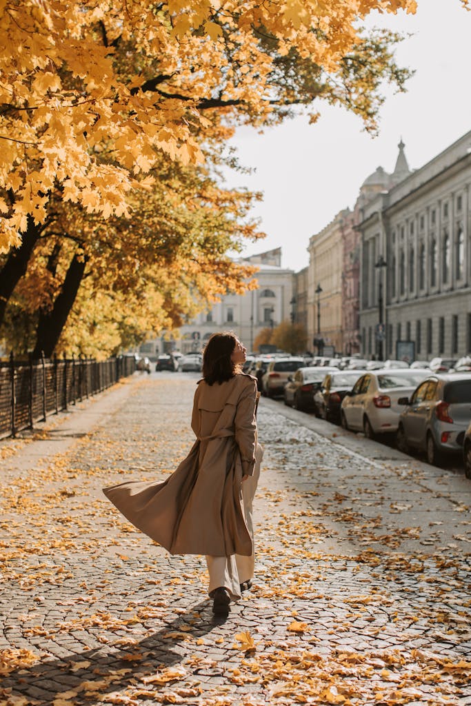 A woman in a trench coat walks down a leaf-covered sidewalk on a sunny autumn day.