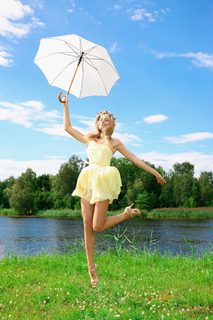 A woman in a yellow dress jumps joyfully with an umbrella by a scenic lakeside on a sunny summer day.