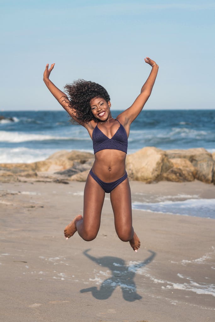 A woman in swimwear joyfully jumps on a sunny beach, exuding happiness.