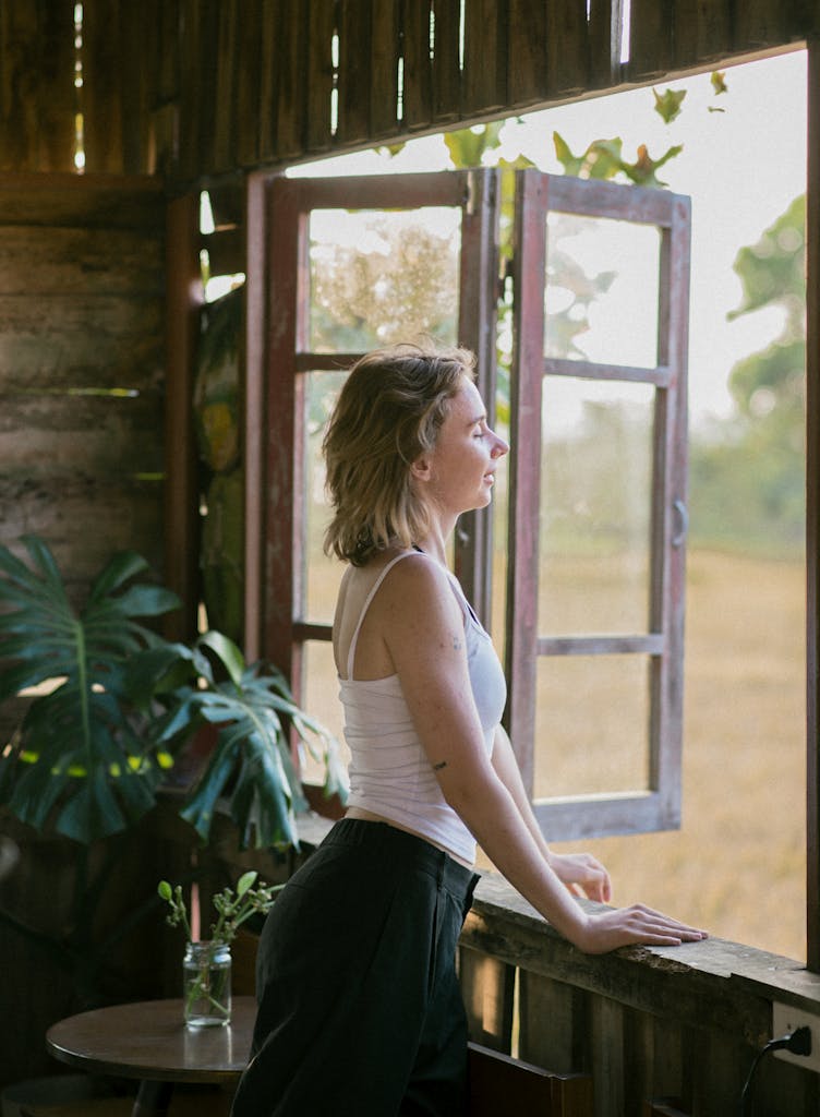 A woman looks out a rustic window at the serene countryside during sunset, capturing peaceful solitude.