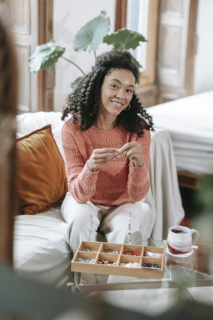 A woman smiling while making handmade jewelry at home with beads on a cozy couch.