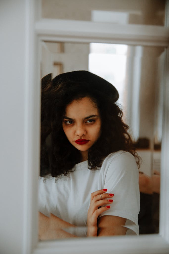 A woman with curly hair looks pensively into a mirror, indoors.