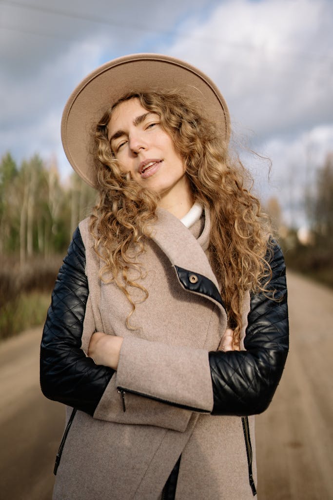 A woman with curly hair wearing a hat poses outdoors in a scenic fall setting.