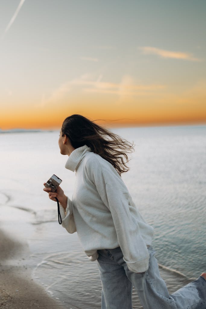 A woman with flowing hair holds a camera by the beach during a serene sunset.