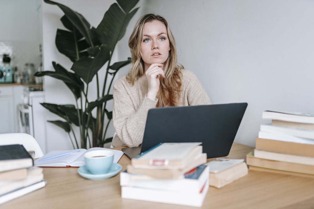 A woman works thoughtfully at a table with a laptop, books, and coffee in a cozy indoor setting.