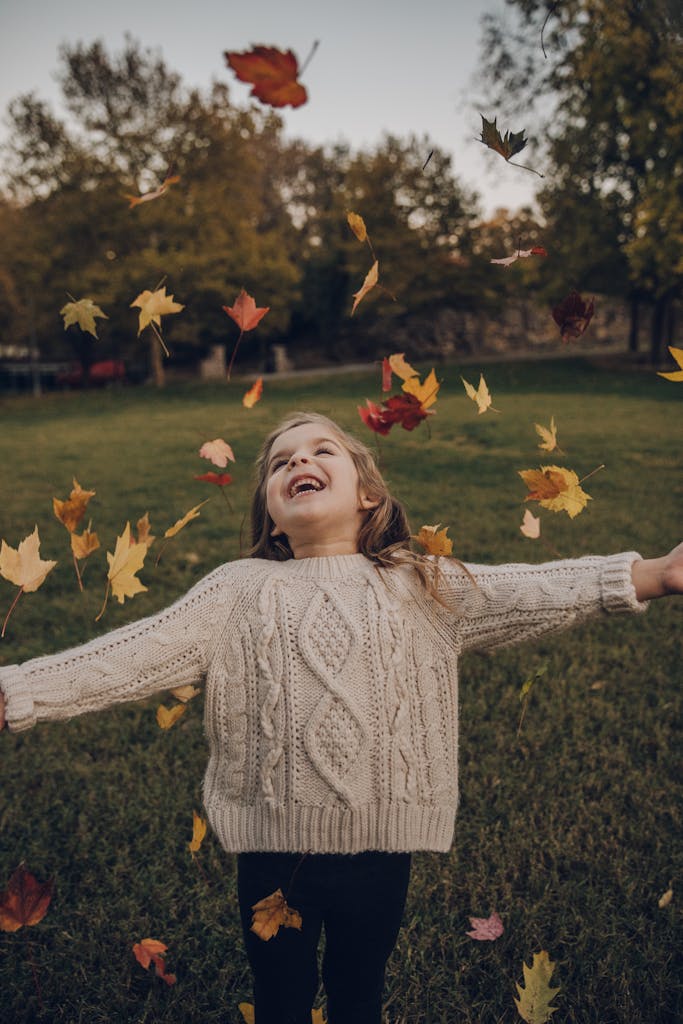A young girl enjoys a playful moment surrounded by vibrant autumn leaves in a park.