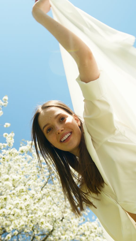 A young woman in a white dress joyfully enjoys springtime outdoors with blooming trees.