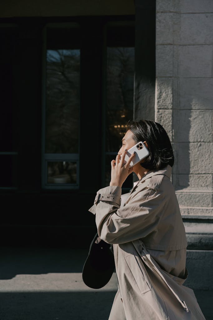 Asian businesswoman walking outdoors in a trench coat and making a phone call.