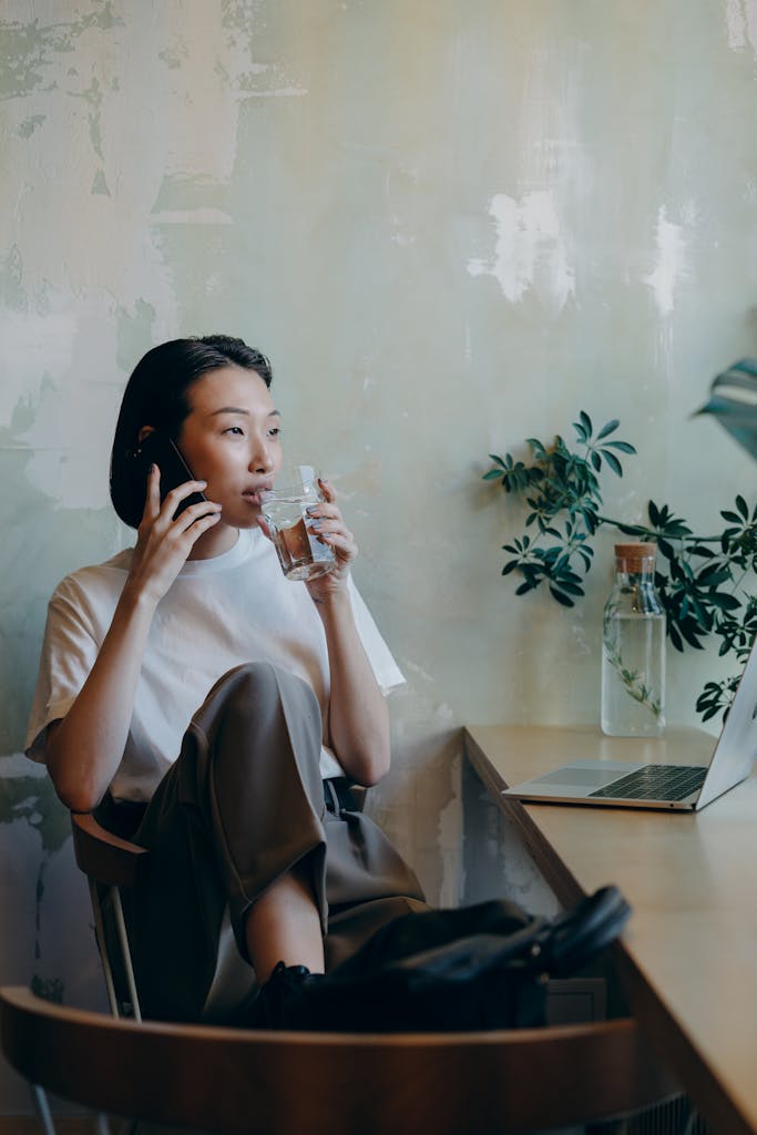 Asian woman multitasking with a phone call and drink in a café setting, wearing a white shirt.