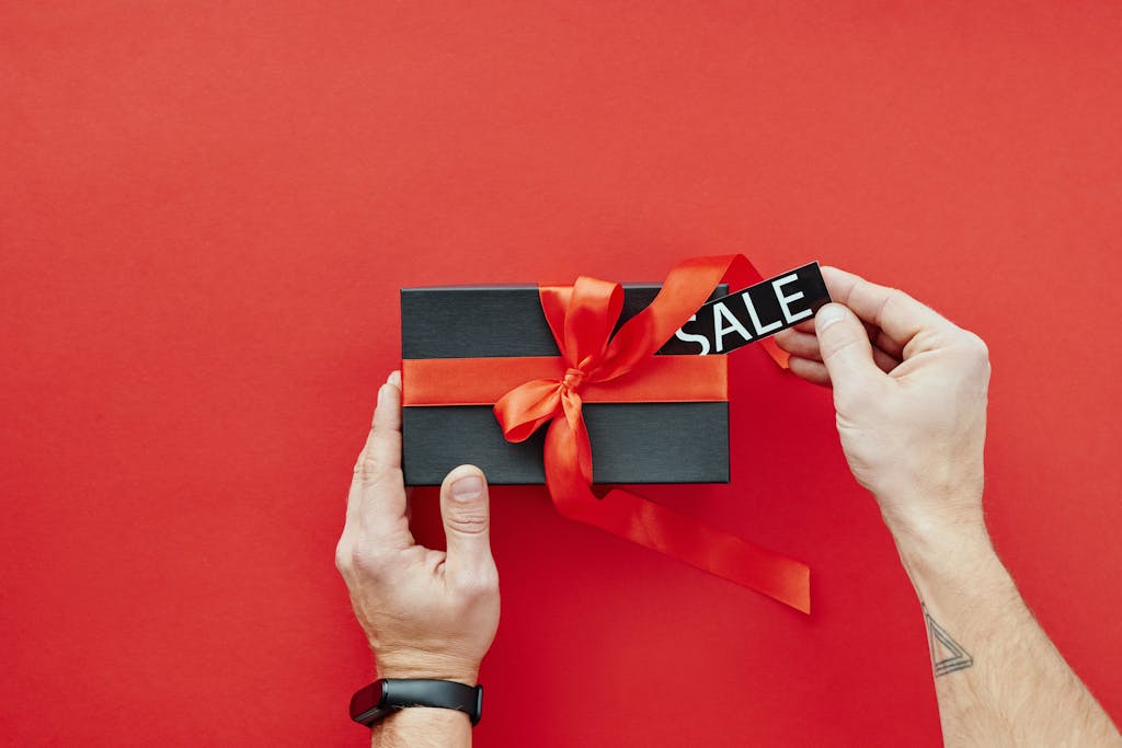 Black gift box with red ribbon and sale tag against a vibrant red background, perfect for promotions.