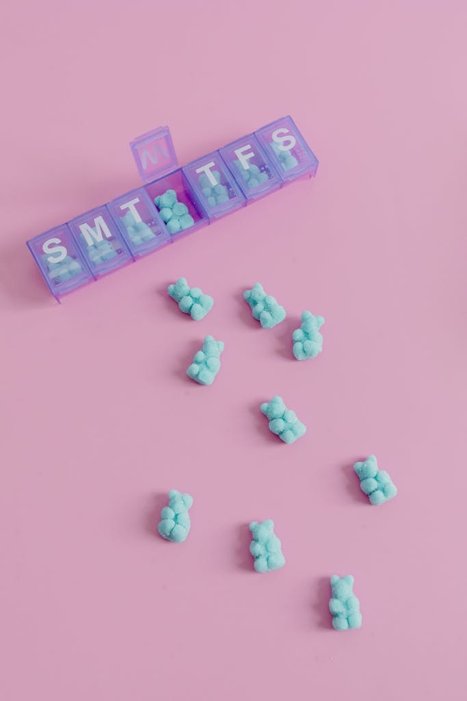 Bright blue gummy bears placed creatively in a weekly pill organizer on a pastel pink background.