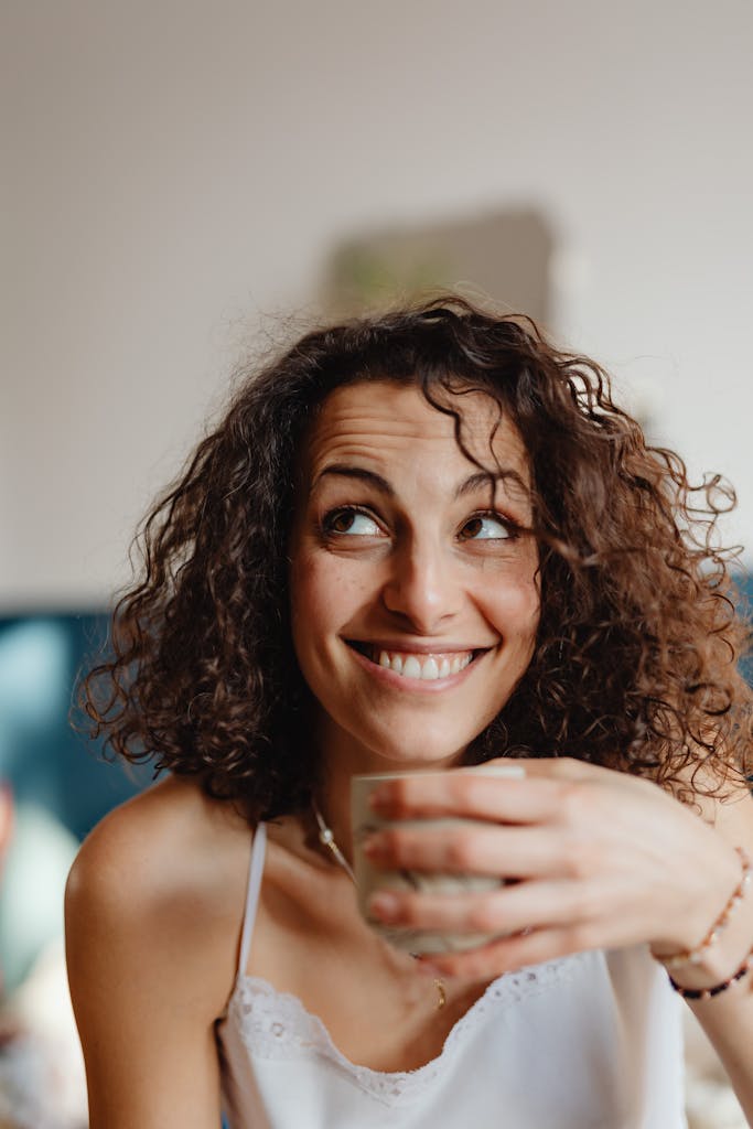 Cheerful woman with curly hair holding a coffee cup and smiling indoors.