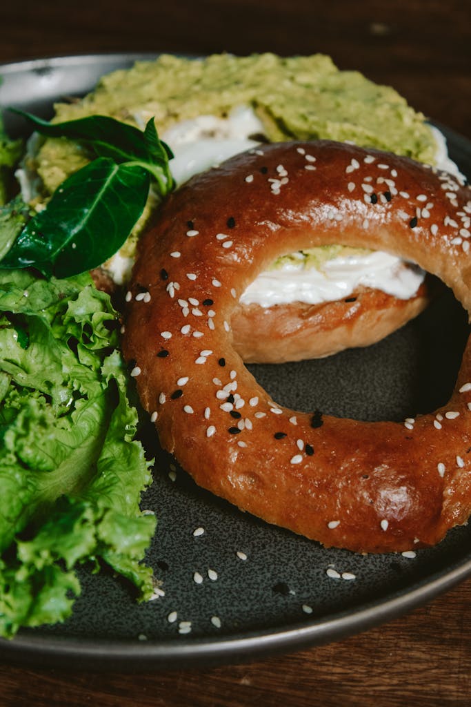 Close-up of a sesame bagel topped with avocado and cream cheese, served with fresh greens.