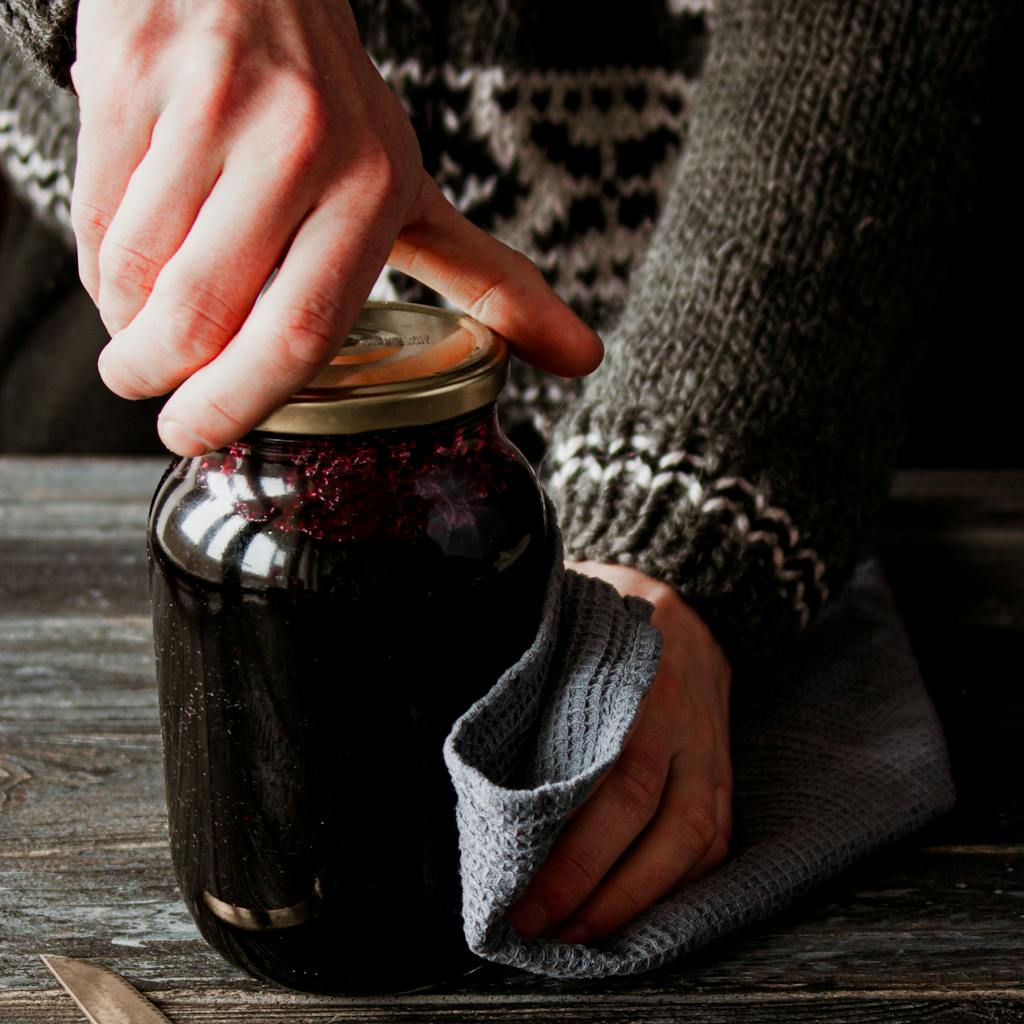 Close-up of hands opening a glass jar on a rustic table, showcasing homemade preserves in cozy kitchen light.