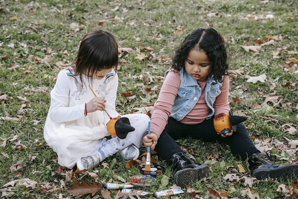 Concentrated little girls painting on pumpkins with brushes together sitting on ground in park