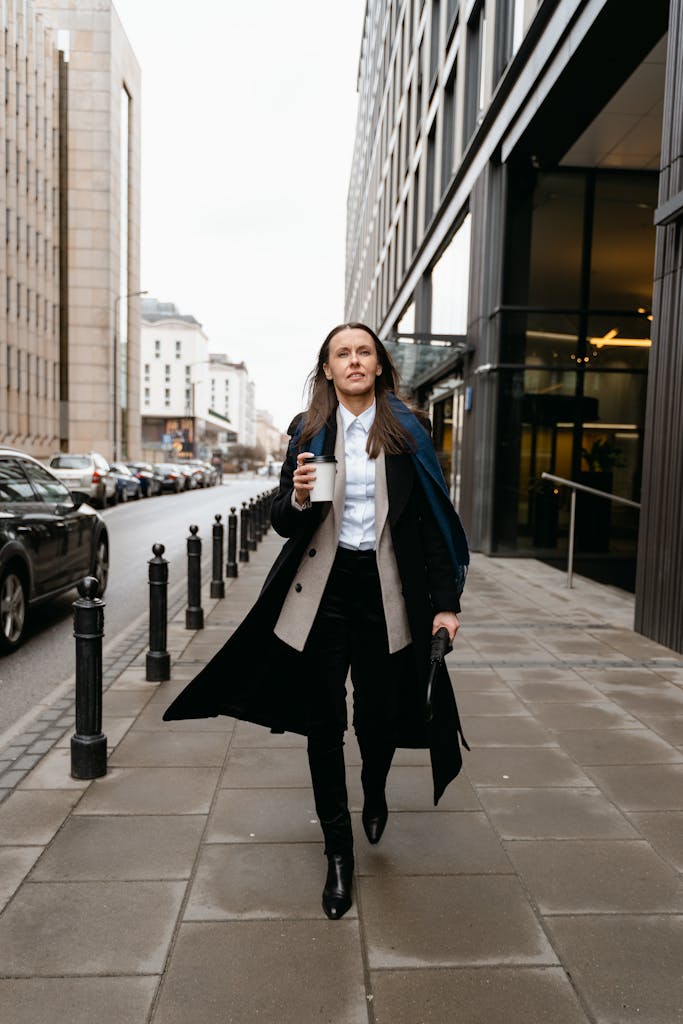 Confident businesswoman walking on city sidewalk holding coffee, dressed in stylish coat.