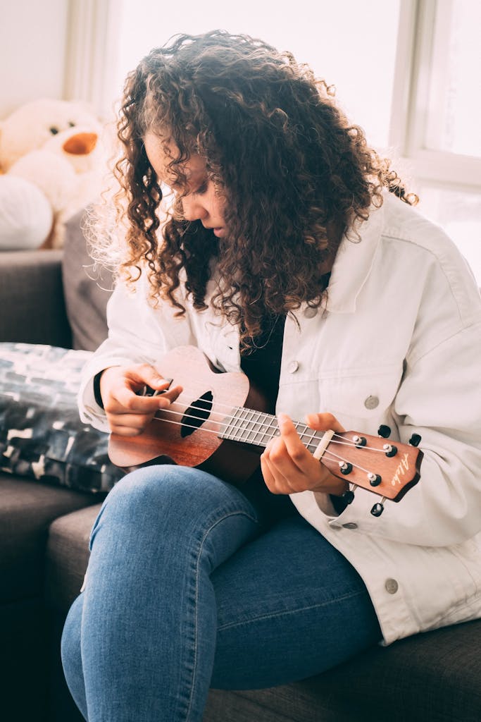 Curly haired woman strumming a ukulele indoors, sitting comfortably on a sofa.