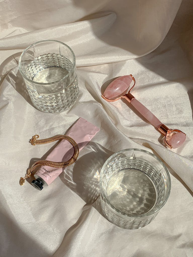 Flat lay of beauty items with rose quartz roller and glass on a satin sheet, emphasizing self-care.