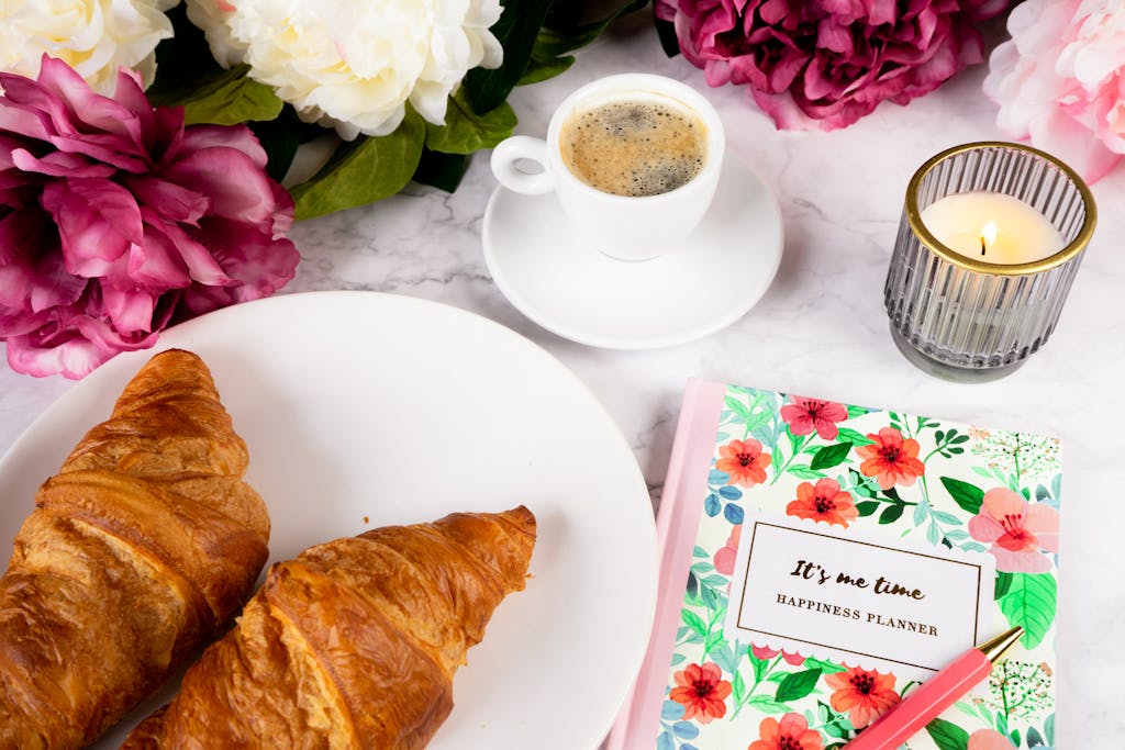 Flat lay of breakfast with croissants, coffee, planner, candle and flowers.